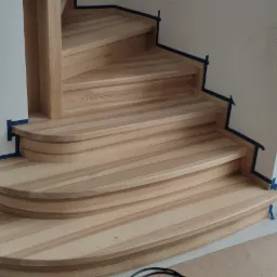 Jasne, drewniane schody z zaokrąglonymi stopniami, zabezpieczone taśmą malarską przed cyklinowaniem, widok z dołu na tle okna z zimowym krajobrazem.