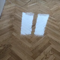 Parkiet ułożony w jodełkę, świeżo polakierowany, z widocznym odbiciem okna i grzejnikiem żeberkowym w tle.