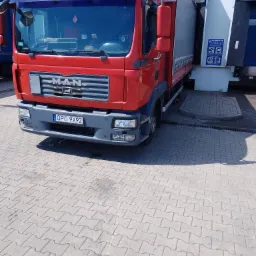 Czerwona ciężar&oacute;wka MAN z naczepą, zaparkowana przy rampie załadunkowej z widocznym logo firmy Wiraz na szybie.