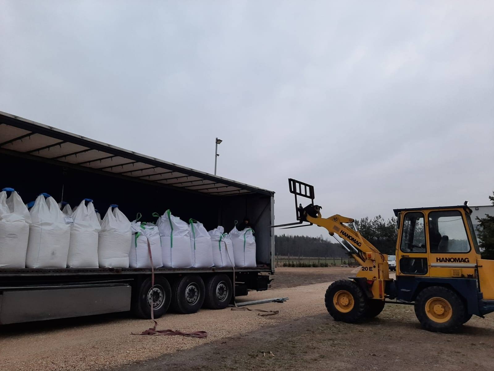 Żółty wózek widłowy Hanomag ładuje białe worki typu big bag z zielonymi uchwytami do naczepy ciężarówki na żwirowym placu, w tle widoczne drzewa.