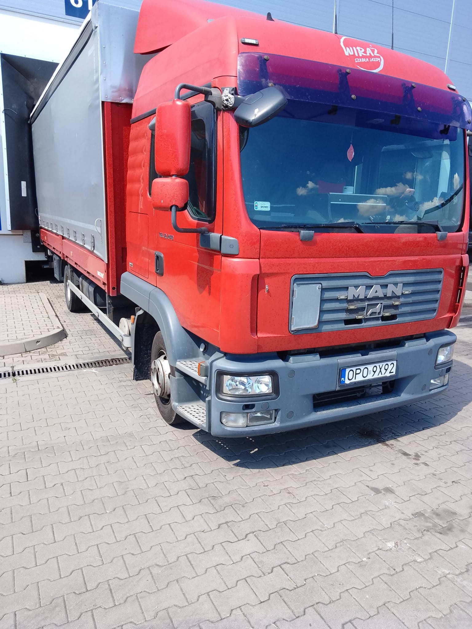Czerwona ciężarówka MAN z naczepą częściowo schowana w doku załadunkowym, widoczny numer rejestracyjny OPO 9X92 i logo firmy Wiraz Szkoła Jazdy na szybie.
