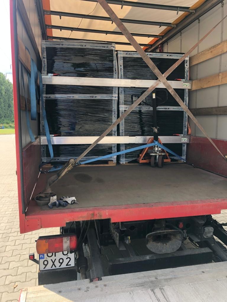 Wnętrze skrzyni ładunkowej samochodu ciężarowego z paletami zabezpieczonymi pasami transportowymi, widoczny wózek paletowy i rękawice robocze.