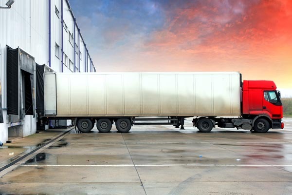 Czerwona ciężarówka z naczepą zaparkowana przy rampie magazynowej, widoczne otwarte drzwi magazynu i mokry asfalt.