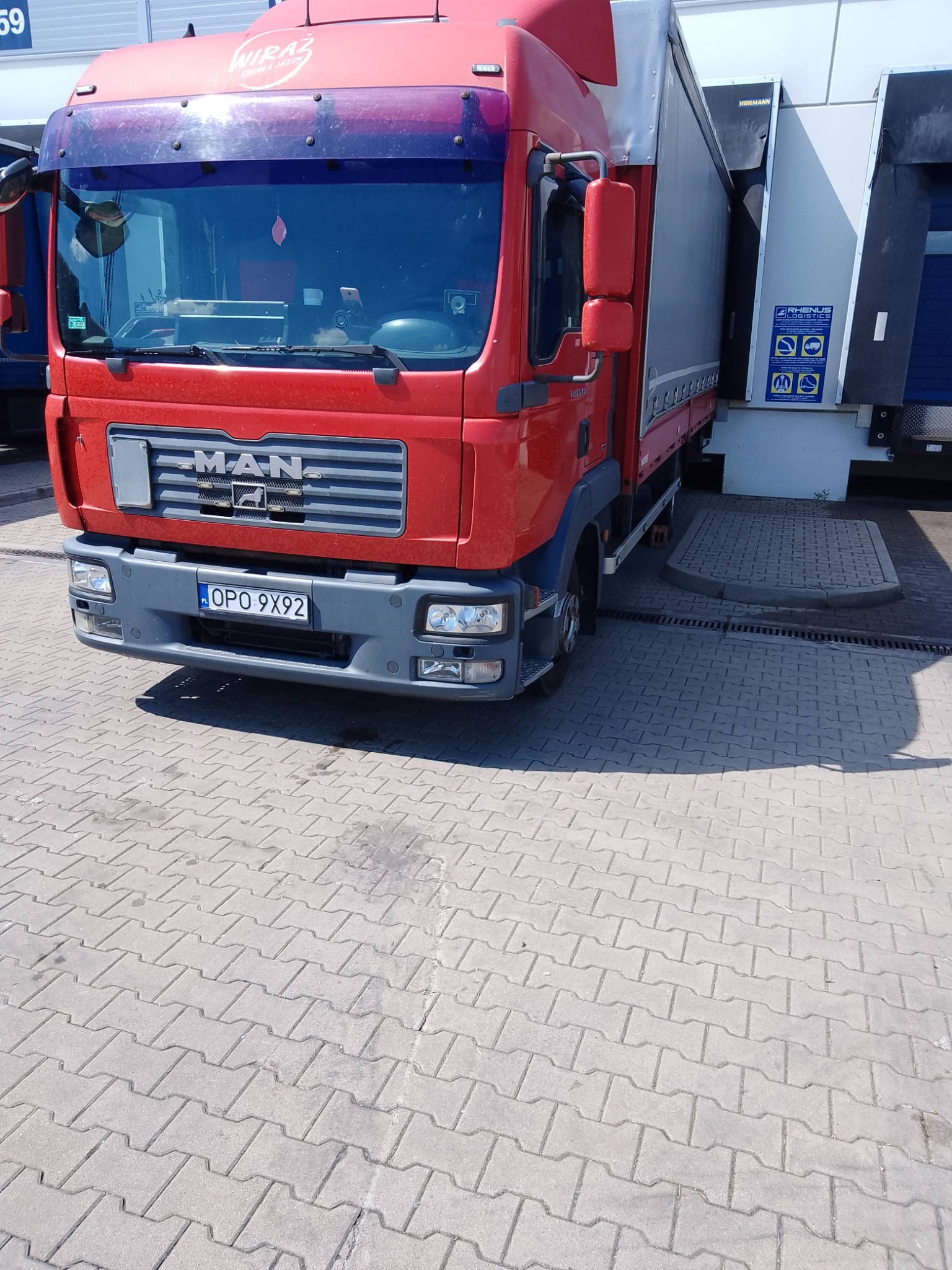 Czerwona ciężarówka MAN z naczepą, zaparkowana przy rampie załadunkowej z widocznym logo firmy Wiraz na szybie.