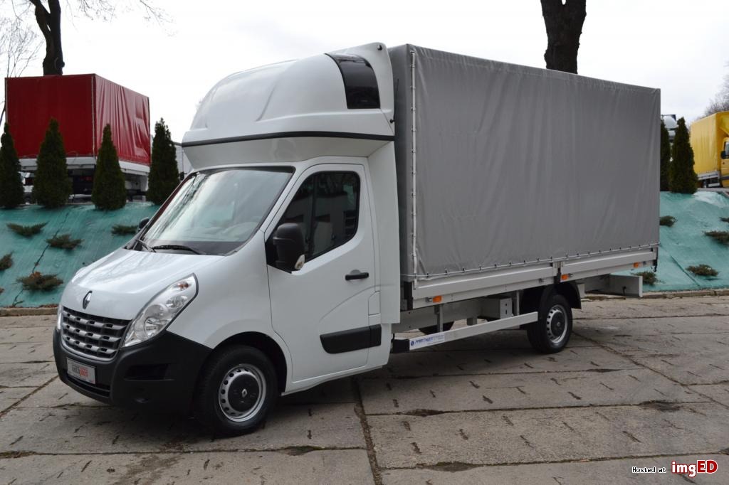 Biały samochód dostawczy Renault Master z plandeką na betonowym podłożu, widok z boku, w tle inne pojazdy i drzewa.