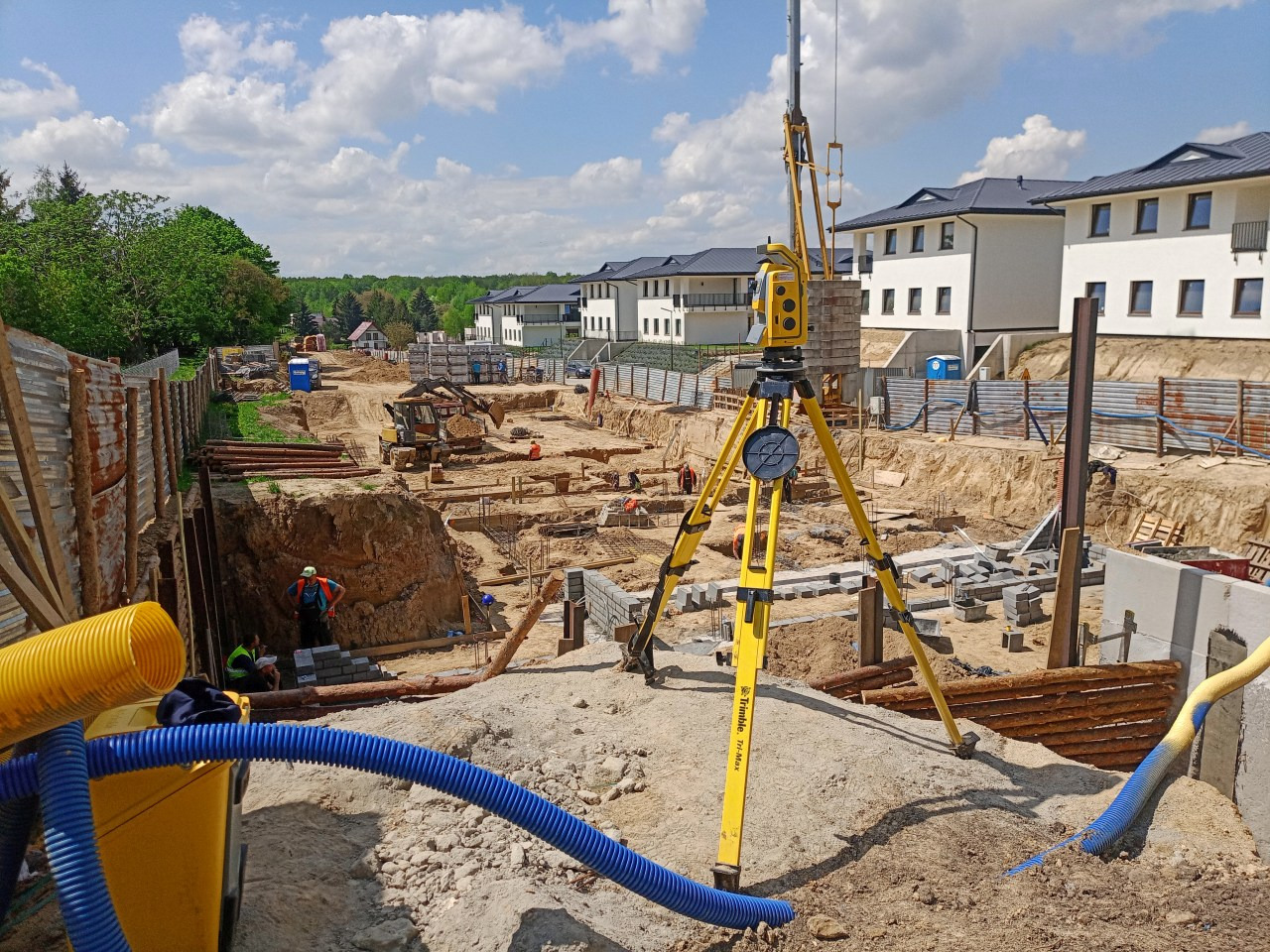 Żółty teodolit Trimble Tri-Max na trójnogu, ustawiony na terenie budowy z wykopami, zbrojeniami i fundamentami pod budynki mieszkalne w tle, błękitne niebo z chmurami.