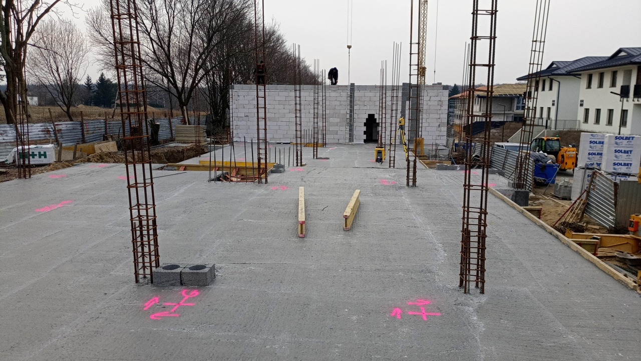 Wylany betonowy fundament z wystającymi prętami zbrojeniowymi, różowe oznaczenia geodezyjne, ściany z betonu komórkowego w tle, widoczni pracownicy na górze ściany, budowa w trakcie.