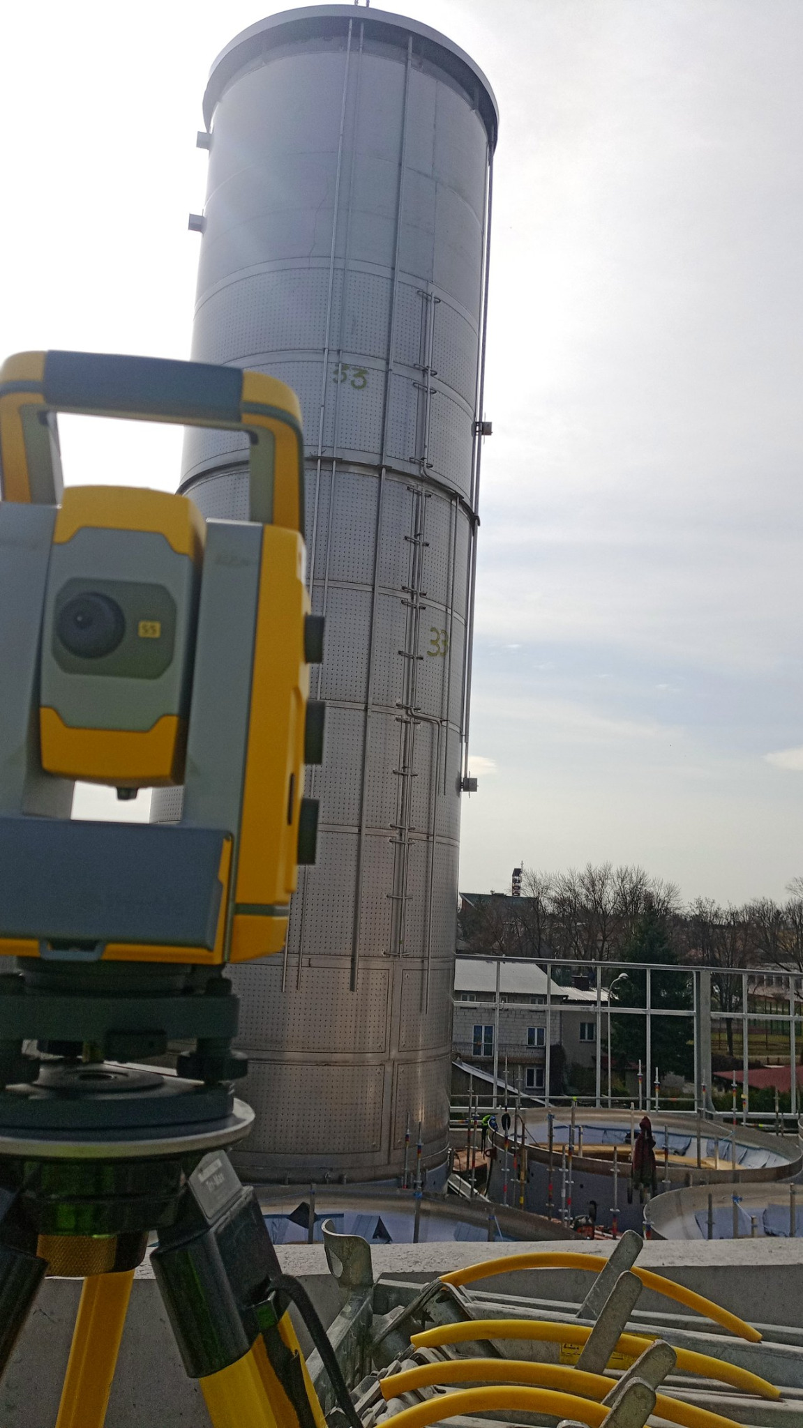 Żółto-szary teodolit na trójnogu, ustawiony na betonowej konstrukcji, w tle wysoki, metalowy silos z numerami 33 i 53, widoczne budynki i drzewa.