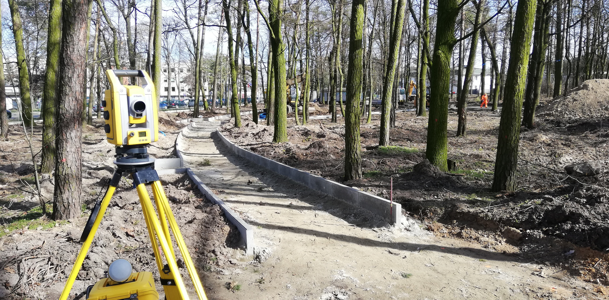 Żółty teodolit na statywie ustawiony przy budowie ścieżki w parku, w tle pracujący sprzęt budowlany i robotnik w pomarańczowym kombinezonie.