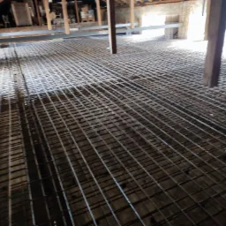 Siatka zbrojeniowa ułożona na podłożu wewnątrz budynku, przygotowana do wylewki betonowej, widoczna drewniana konstrukcja stropu.