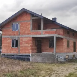 Dwukondygnacyjny dom w trakcie budowy z czerwonej cegły, z widocznymi oknami i betonowymi elementami konstrukcyjnymi, na tle pochmurnego nieba i drzew.