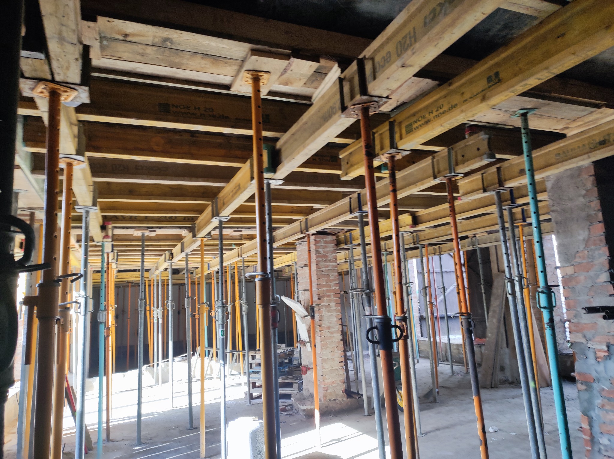 Konstrukcja szalunkowa stropu podparta stalowymi stemplami w surowym wnętrzu budynku w trakcie budowy, widoczne fragmenty ceglanej ściany w tle.