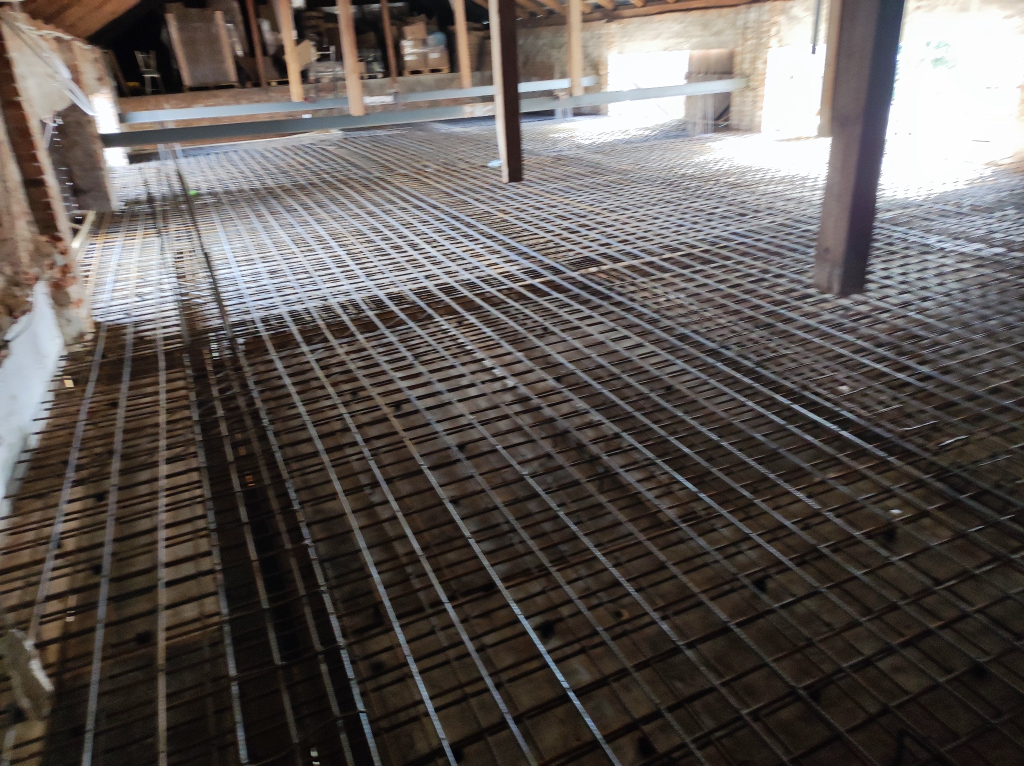 Siatka zbrojeniowa ułożona na podłożu wewnątrz budynku, przygotowana do wylewki betonowej, widoczna drewniana konstrukcja stropu.