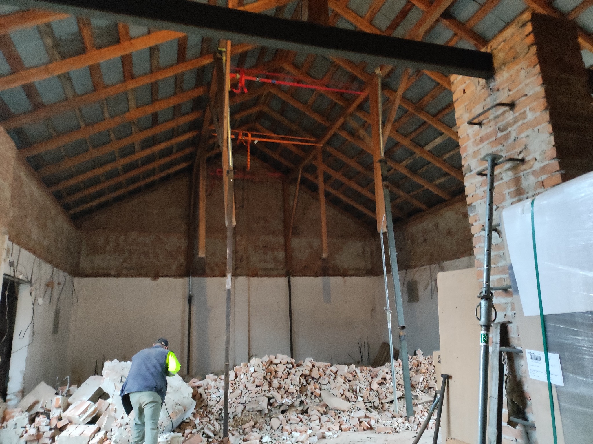Wnętrze remontowanego domu z widocznym drewnianym dachem podpartym metalowymi stemplami, gruz ceglany na podłodze i pracownik w kamizelce odblaskowej