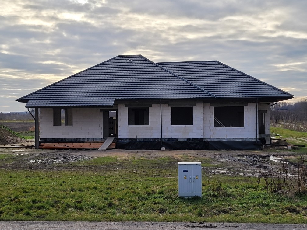 Niedokończony dom jednorodzinny z szarej dachówki, widoczne bloczki betonowe, otwory okienne bez ram, drewniane deski na ziemi, szara skrzynka elektryczna na pierwszym planie, pochmurne niebo w tle.