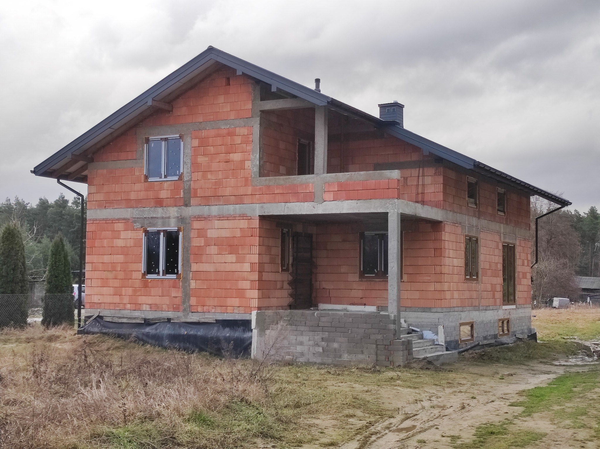Dwukondygnacyjny dom w trakcie budowy z czerwonej cegły, z widocznymi oknami i betonowymi elementami konstrukcyjnymi, na tle pochmurnego nieba i drzew.