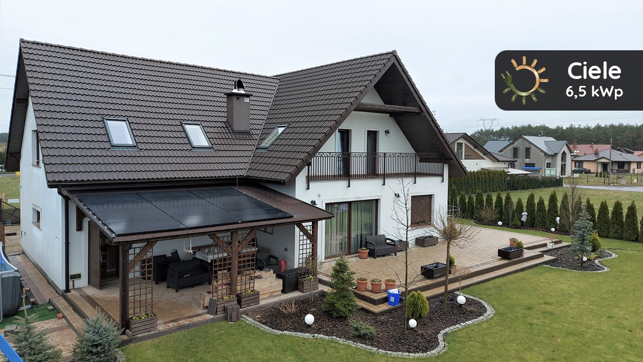 Nowoczesny dom jednorodzinny z brązowym dachem i panelami fotowoltaicznymi na zadaszeniu tarasu, widok z ogrodu z dekoracyjnymi krzewami i kulistymi lampami, logo firmy Ciele 6,5 kWp w prawym...