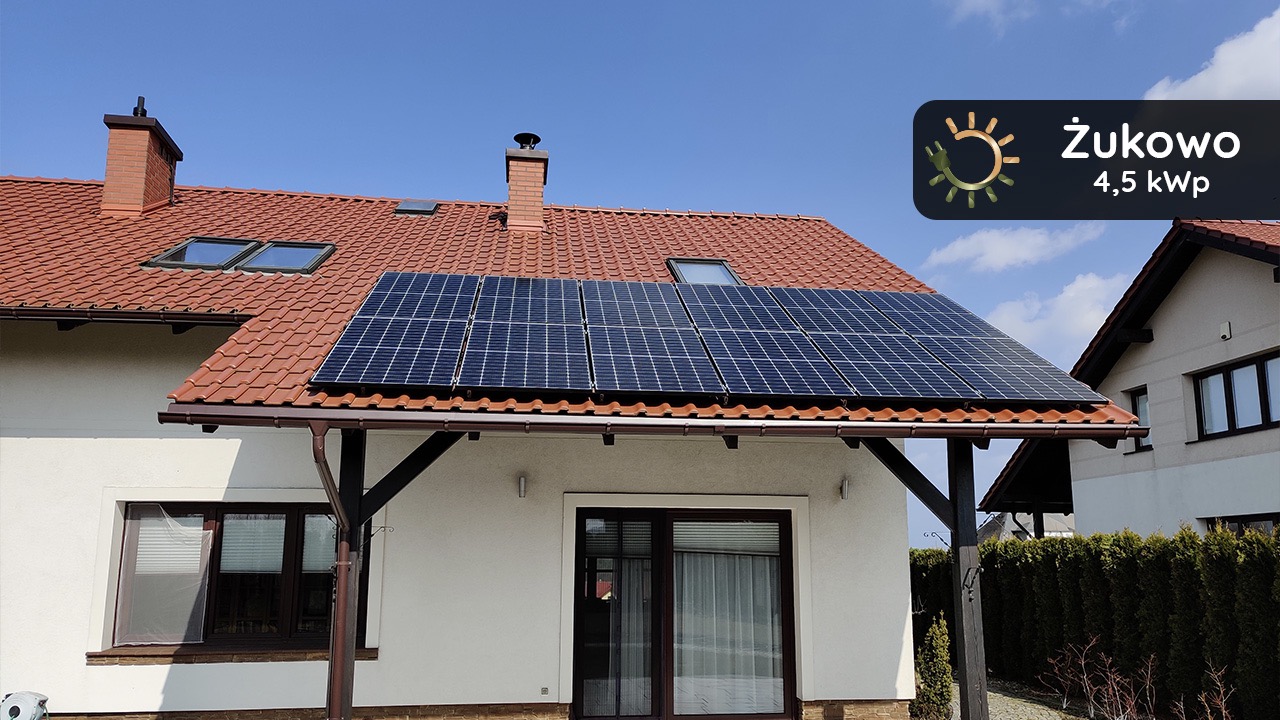 Panel słoneczny na dachu domu jednorodzinnego z czerwoną dachówką, widoczne okna dachowe i komin, błękitne niebo w tle, logo firmy w prawym górnym rogu.