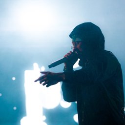 Wideofilmowanie - Adam Miłaszewski - Artysta w ciemnej bluzie z kapturem i okularach, wykonujący utwór do mikrofonu na scenie. Dynamiczne ujęcie z boku, z mocnym, jasnym podświetleniem i rozmytymi światłami w tle, tworzącymi mglistą...