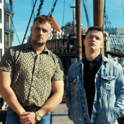 Dwaj mężczyźni pozują na tle żaglowca i zabudowy miejskiej, prawdopodobnie podczas kręcenia teledysku.