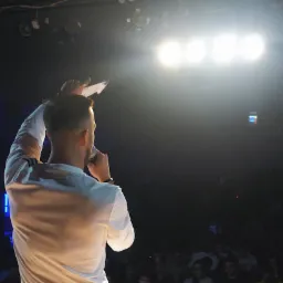 Prezenter z mikrofonem na scenie podczas Mistrzostw Polski Freestyle Football 2019, oświetlony reflektorami, z publicznością w tle.