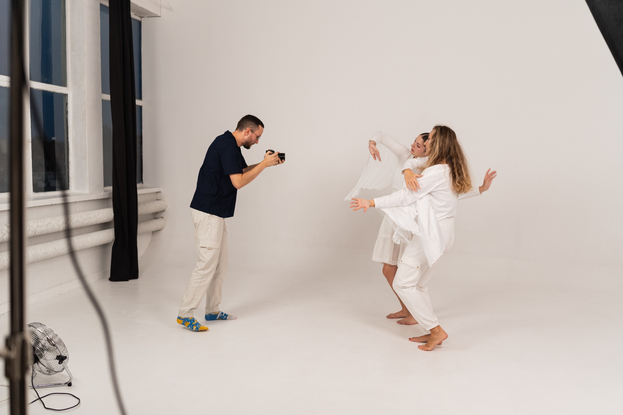 Fotograf robi zdjęcie dwóm modelkom w białych strojach w studiu; widoczny wentylator, okno i kolorowe skarpetki fotografa.