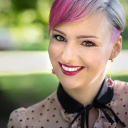 Uśmiechnięta kobieta z kr&oacute;tkimi włosami w odcieniach szarości i r&oacute;żu, z ciemną szminką i kolczykami w kształcie kwiat&oacute;w, ubrana w bluzkę w serduszka na tle zieleni.