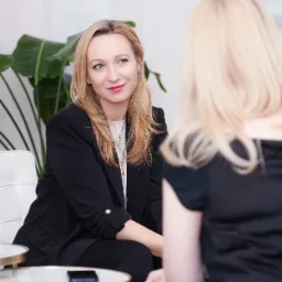 Kobieta w czarnej marynarce uśmiecha się do blondynki podczas sesji terapeutycznej w jasnym pokoju z roślinami w tle.