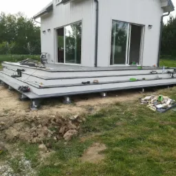 Budowa tarasu przy białym domu z dużymi oknami przesuwnymi; widoczne narzędzia, elementy konstrukcyjne i wykopana ziemia.