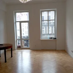 Pusty pokój z parkietem ułożonym w jodełkę, białymi ścianami, oknami z widokiem na budynki, i kryształowym żyrandolem.