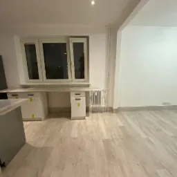 Puste, jasne pomieszczenie po remoncie z nową podłogą panelową, białymi szafkami pod oknem i grzejnikiem, widok na ścianę z wykończonymi listwami przypodłogowymi.