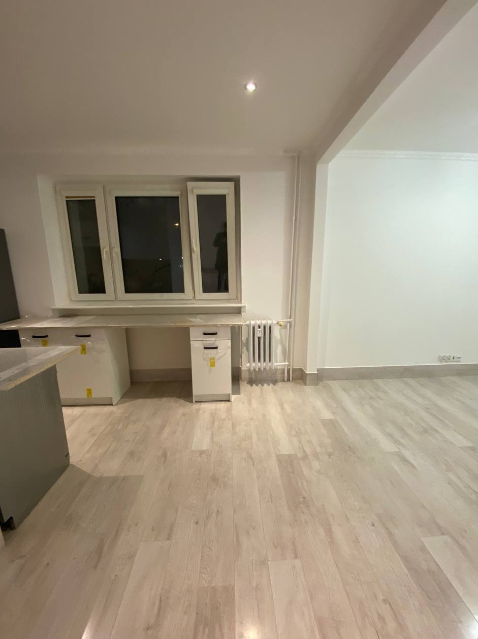 Puste, jasne pomieszczenie po remoncie z nową podłogą panelową, białymi szafkami pod oknem i grzejnikiem, widok na ścianę z wykończonymi listwami przypodłogowymi.