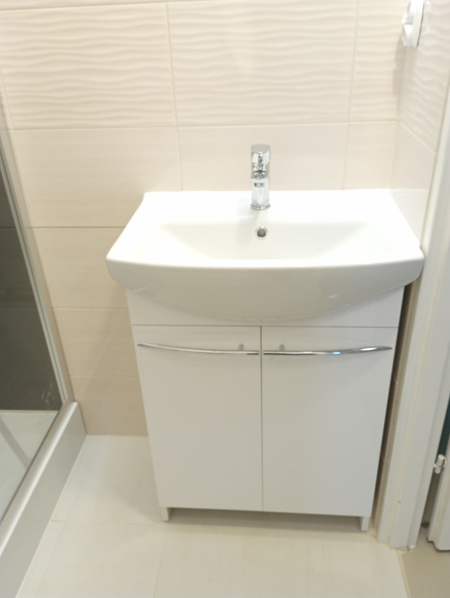 Biała umywalka z szafką w łazience, chromowana bateria, beżowe płytki z delikatnym wzorem, widoczny fragment kabiny prysznicowej.