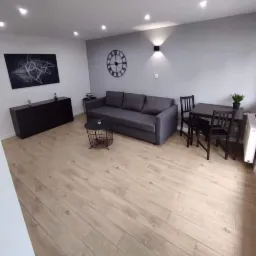 Jasne panele podłogowe w nowoczesnym salonie z szarą sofą, czarną komodą i okrągłym zegarem na ścianie.