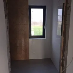 Wnętrze łazienki w trakcie remontu, z ułożonymi brązowymi płytkami z wzorem jodełki na jednej ścianie, białymi płytkami na pozostałych ścianach, czarnym oknem z widokiem na zewnątrz oraz szarą...