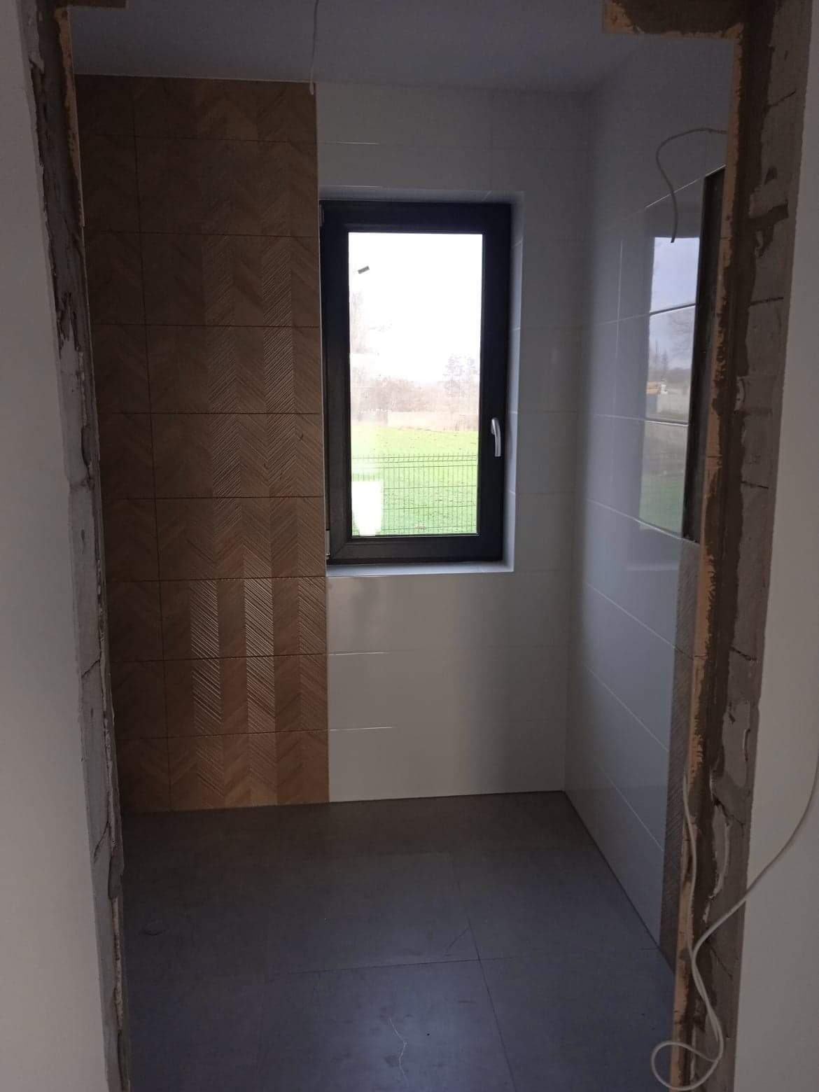 Wnętrze łazienki w trakcie prac wykończeniowych; częściowo ułożone płytki ścienne w dwóch kolorach i wzorach, widoczne surowe obramowanie drzwi i okno z widokiem na zewnątrz.
