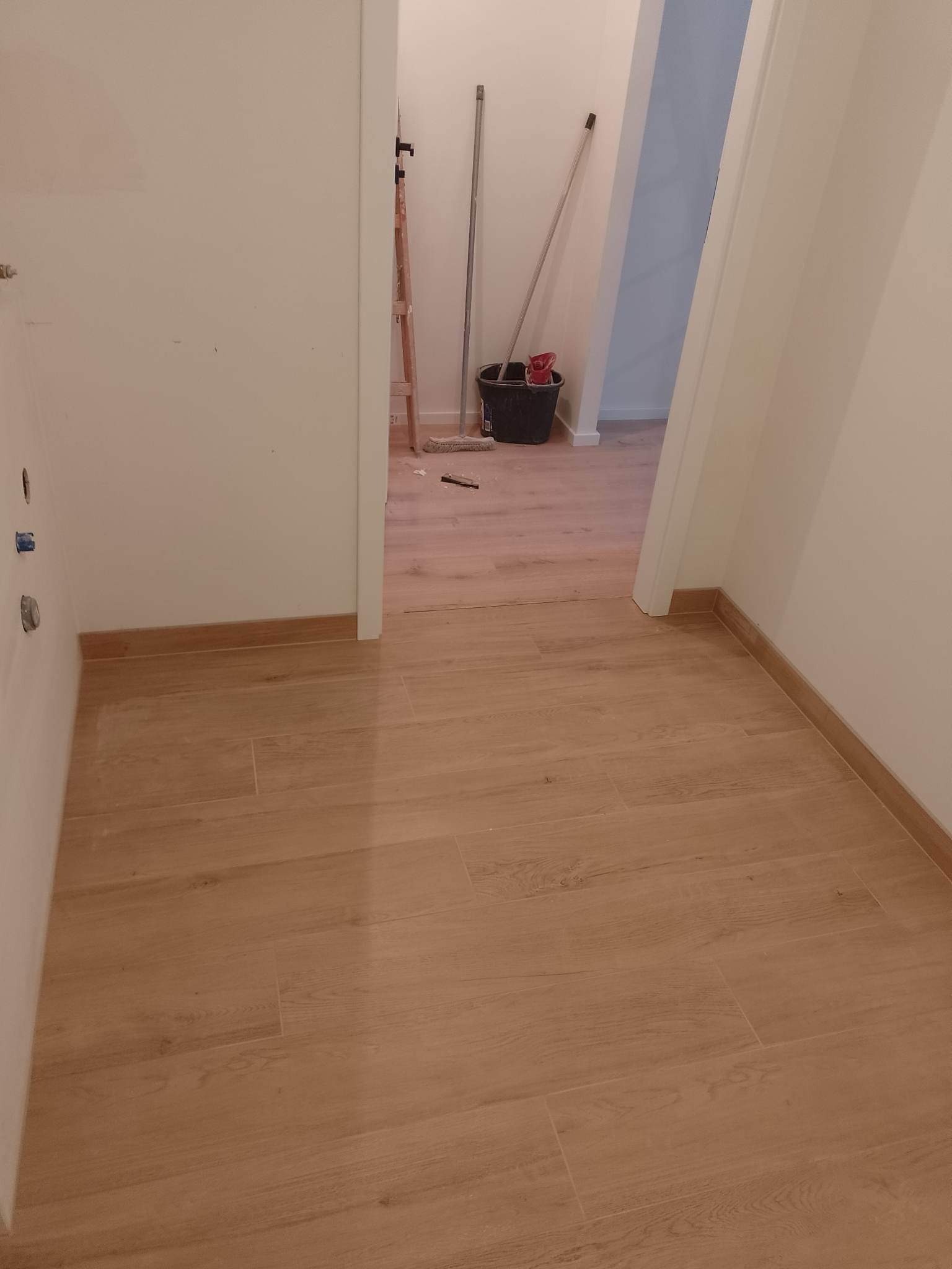 Ułożone płytki podłogowe imitujące drewno w korytarzu, widoczne wiadro, drabina i mop w tle.