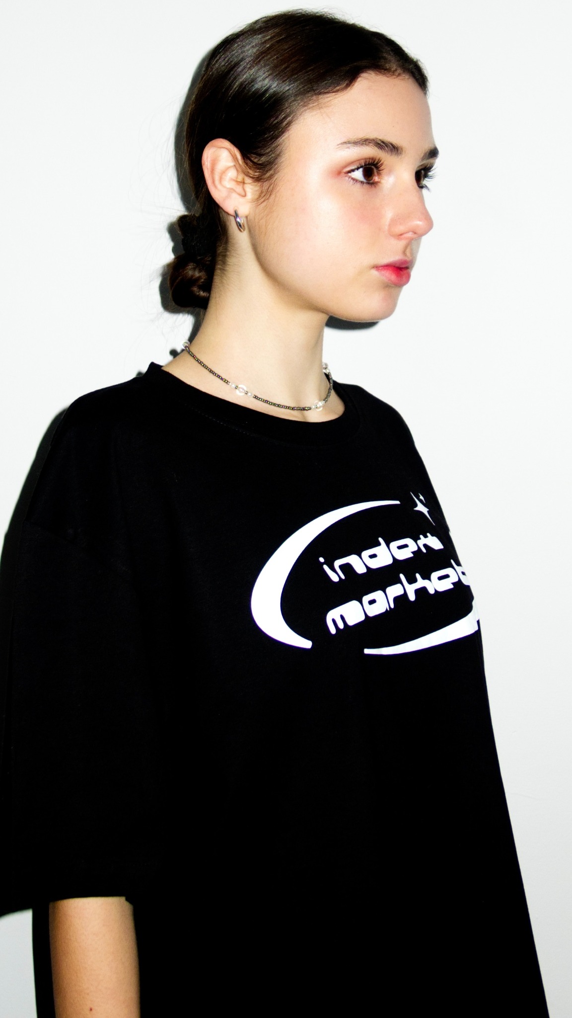 Portret młodej kobiety w czarnej bluzie z białym logo 'index market', srebrny naszyjnik i kolczyki, gładko zaczesane włosy, profil na białym tle.