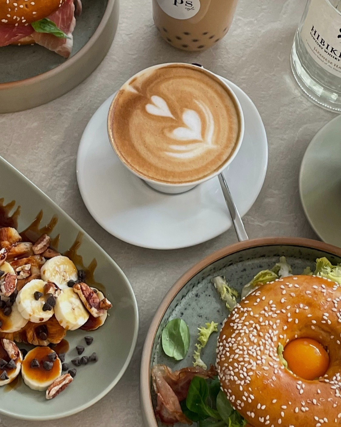 Stół z różnorodnymi daniami: cappuccino z wzorem serca, deser z bananami i czekoladą, bajgiel z jajkiem oraz napój bąbelkowy i butelka wody.