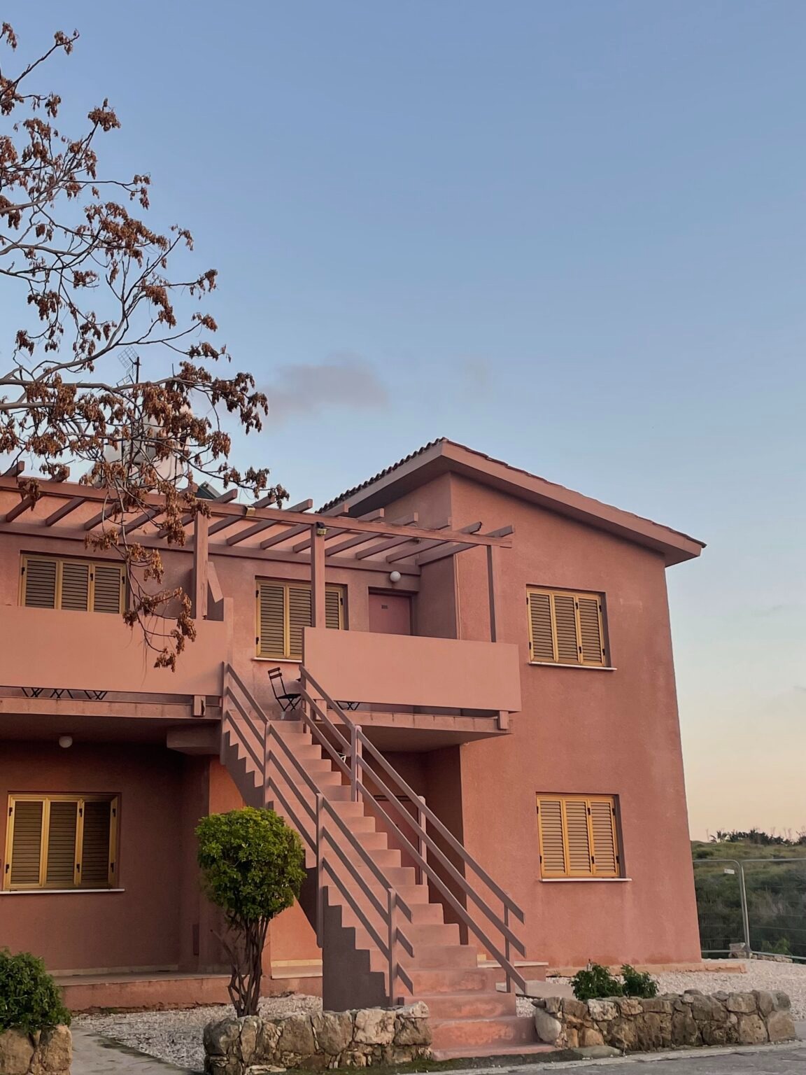 Dwupiętrowy budynek w odcieniach różu z zewnętrznymi schodami, żółtymi okiennicami i minimalistycznym balkonem na tle bladego nieba o zachodzie słońca.