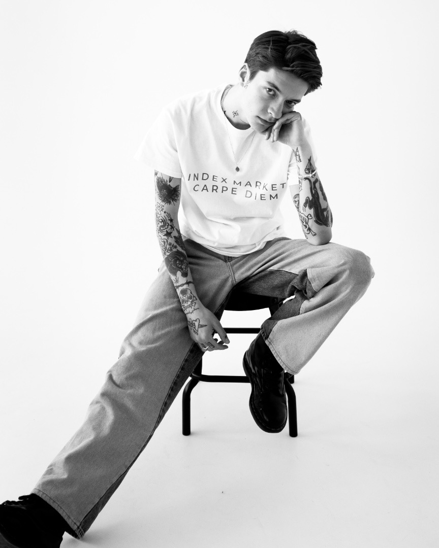 Młody wytatuowany mężczyzna w dżinsach siedzi na czarnym stołku na białym tle, ubrany w białą koszulkę z napisem 'Index Market Carpe Diem'.