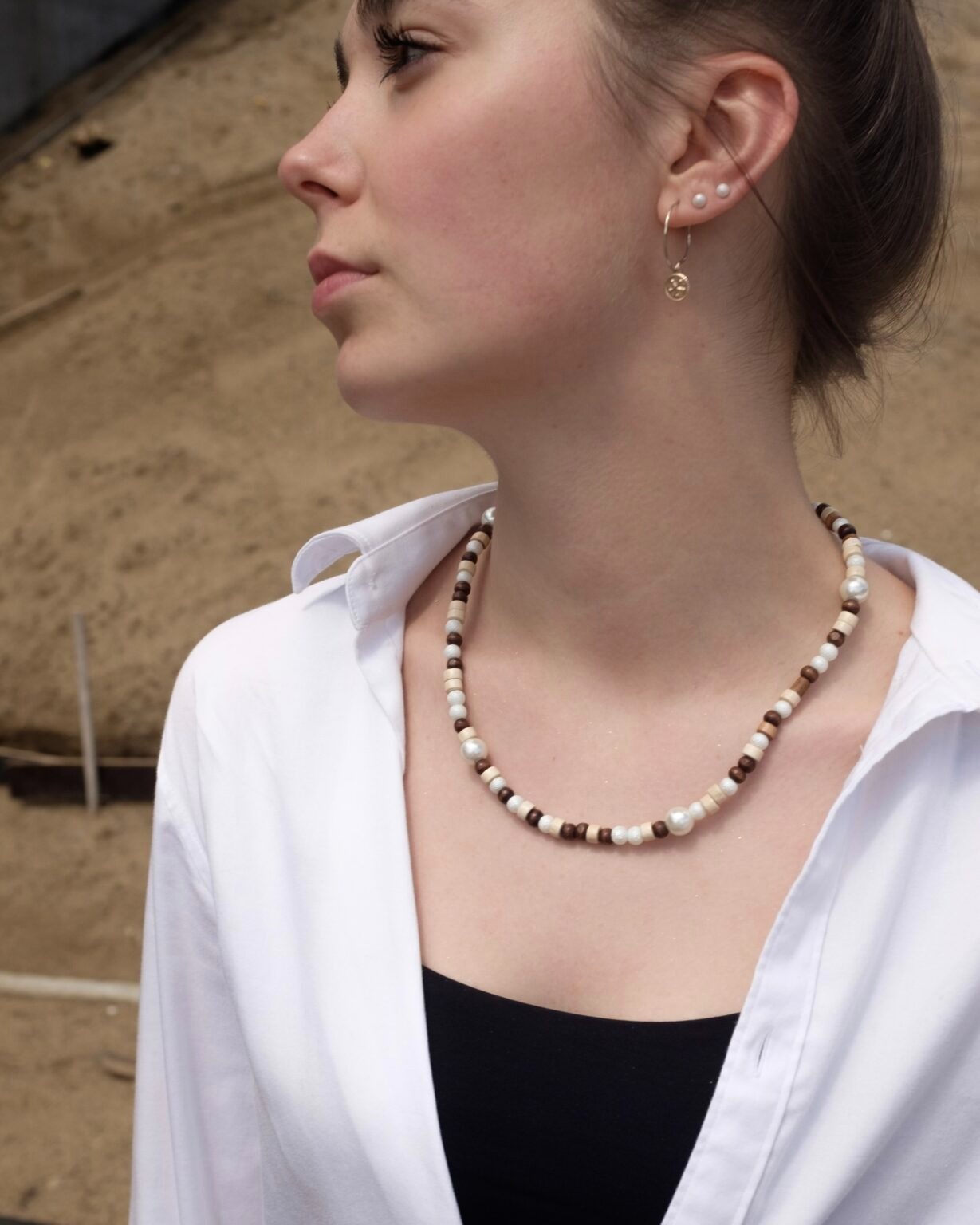 Portret młodej kobiety z naszyjnikiem z drewnianych i perłowych koralików, w białej koszuli i czarnym topie, na tle piaszczystego terenu.