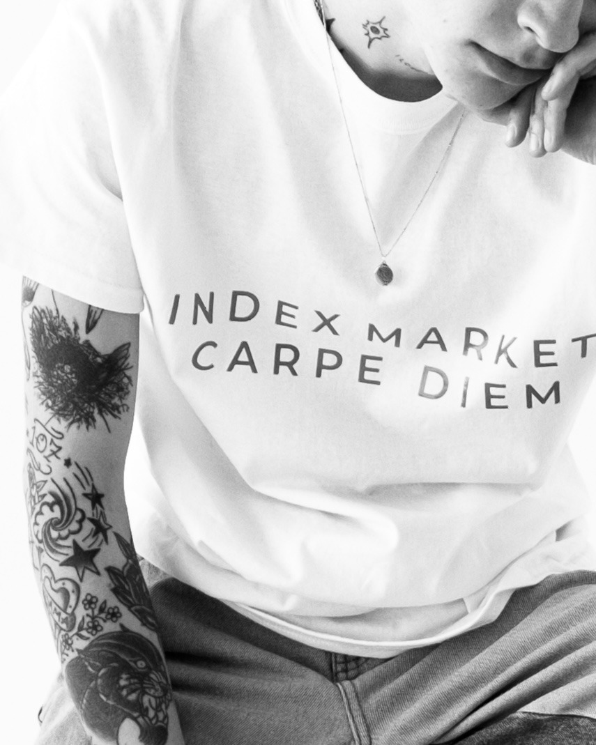 Osoba z tatuażami na ramieniu i szyi, ubrana w białą koszulkę z napisem 'INDEX MARKET CARPE DIEM' i dżinsy, pozuje w czarno-białym ujęciu.