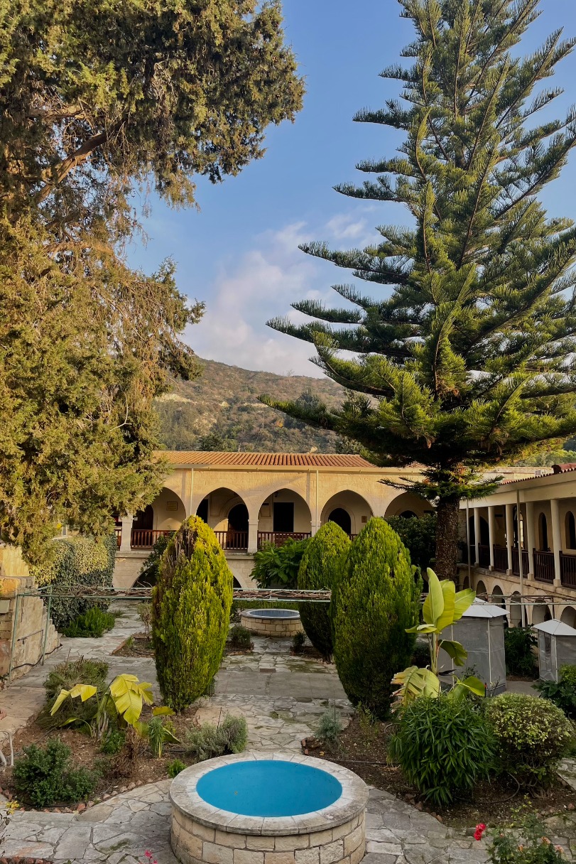 Dziedziniec klasztorny z kamiennymi ścieżkami, studniami z niebieską wodą i bujną roślinnością, otoczony budynkiem z arkadami i widokiem na wzgórza.