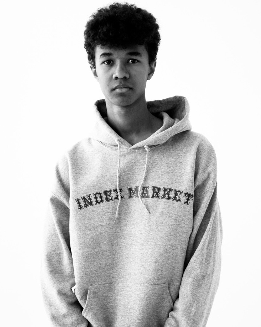 Portret młodego mężczyzny z afro w szarej bluzie z kapturem i napisem 'INDEX MARKET', na białym tle.