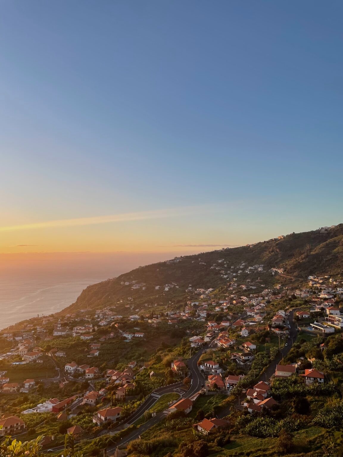 Widok z góry na osiedle domów z czerwonymi dachami na zboczu góry, z oceanem i zachodzącym słońcem w tle.