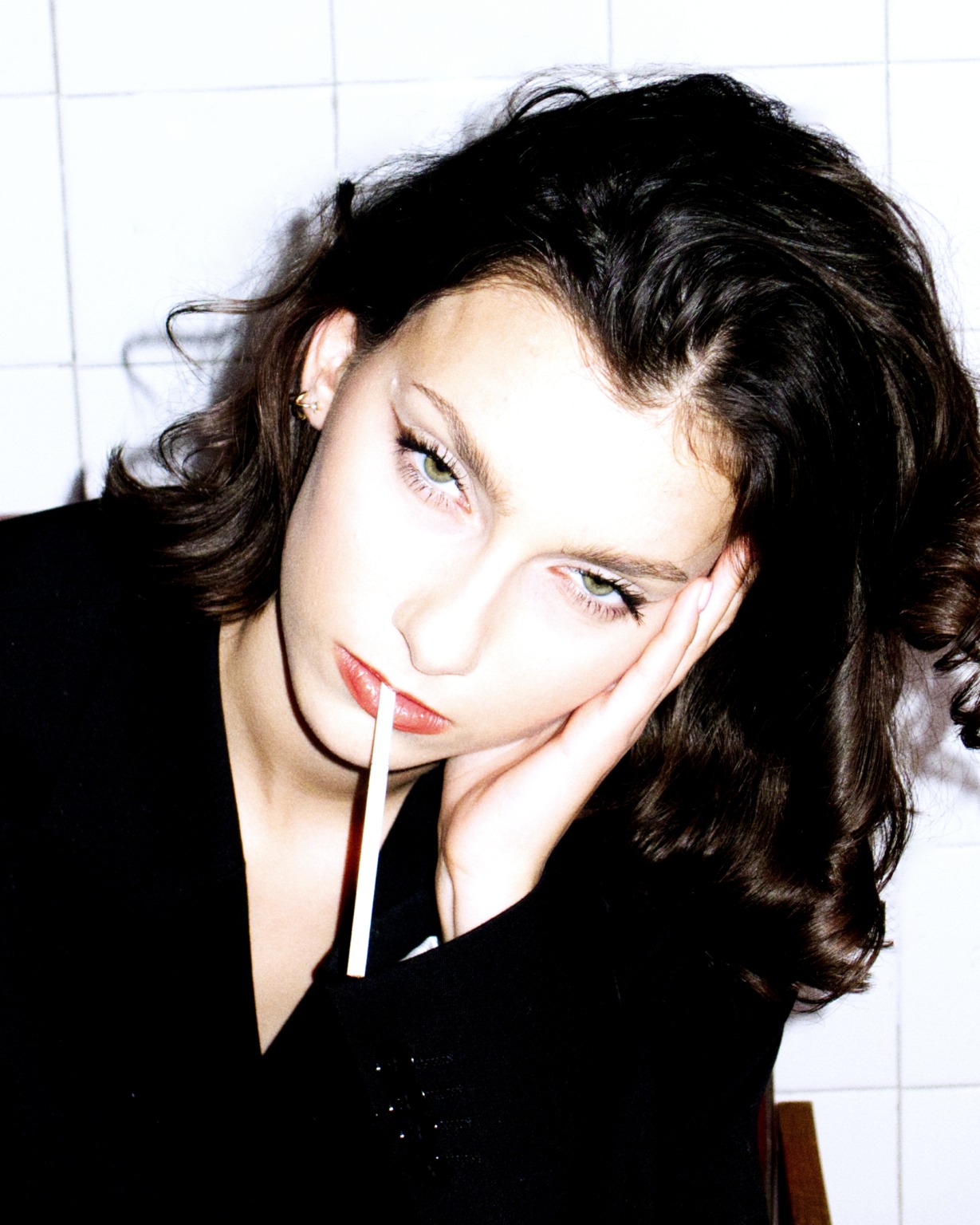 Portret młodej kobiety z ciemnymi włosami i wyrazistym makijażem w Gdyni, trzymającej w ustach białą pałeczkę, opierającej głowę na dłoni na tle białych kafelków.