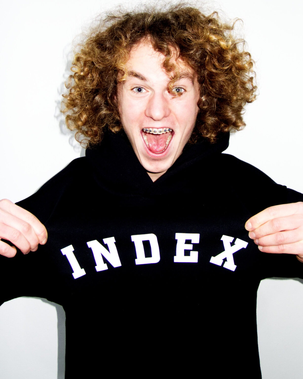 Młody mężczyzna z kręconymi włosami i aparatem ortodontycznym, ubrany w czarną bluzę z napisem 'INDEX', szeroko uśmiechnięty na białym tle.