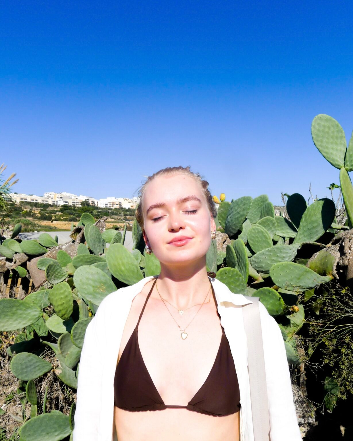 Młoda kobieta w bikini i białej koszuli, z zamkniętymi oczami, stojąca na tle kaktusów i błękitnego nieba w słoneczny dzień.