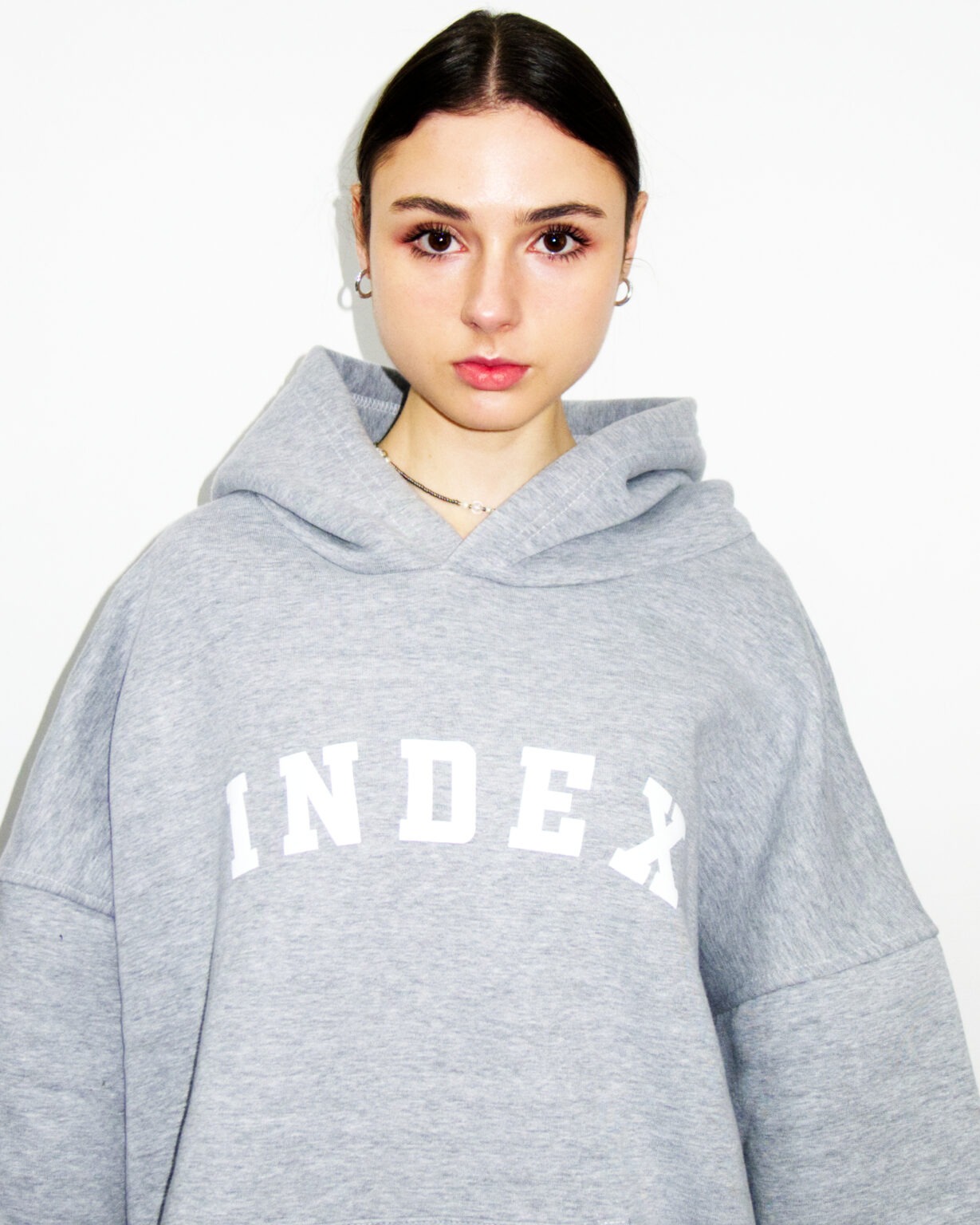 Portret młodej kobiety z ciemnymi włosami, ubrana w szarą bluzę z kapturem z białym napisem 'INDEX', na białym tle.